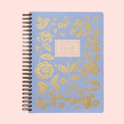 A light blue planner with gold foiled rose detailing, spiral bound and text in the center that reads, "Rifle Paper Co 24-25 17 Month Planner". 