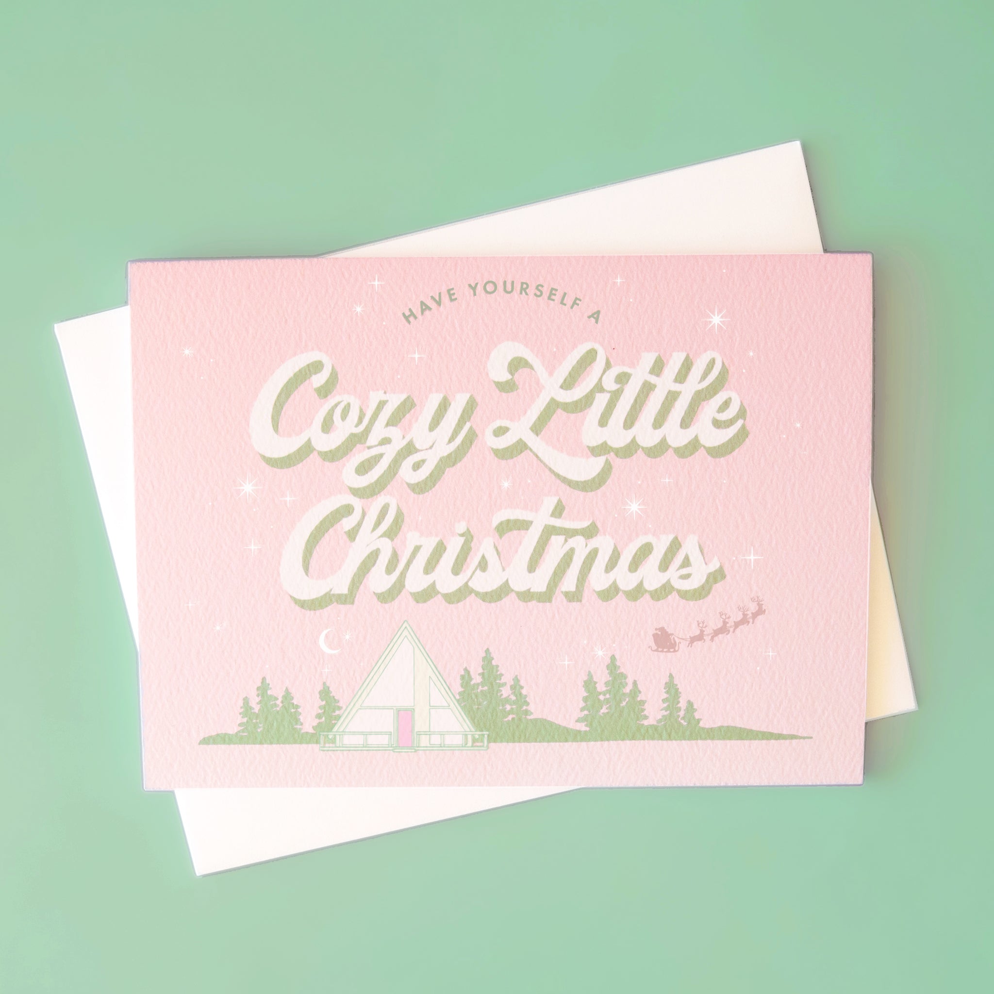 A pink and mint green Christmas card with an A-frame graphic and text above that reads, &quot;Have Yourself A Cozy Little Christmas&quot;.