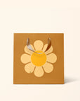 On a white background is a brown gift bag with a large yellow daisy in the center. 