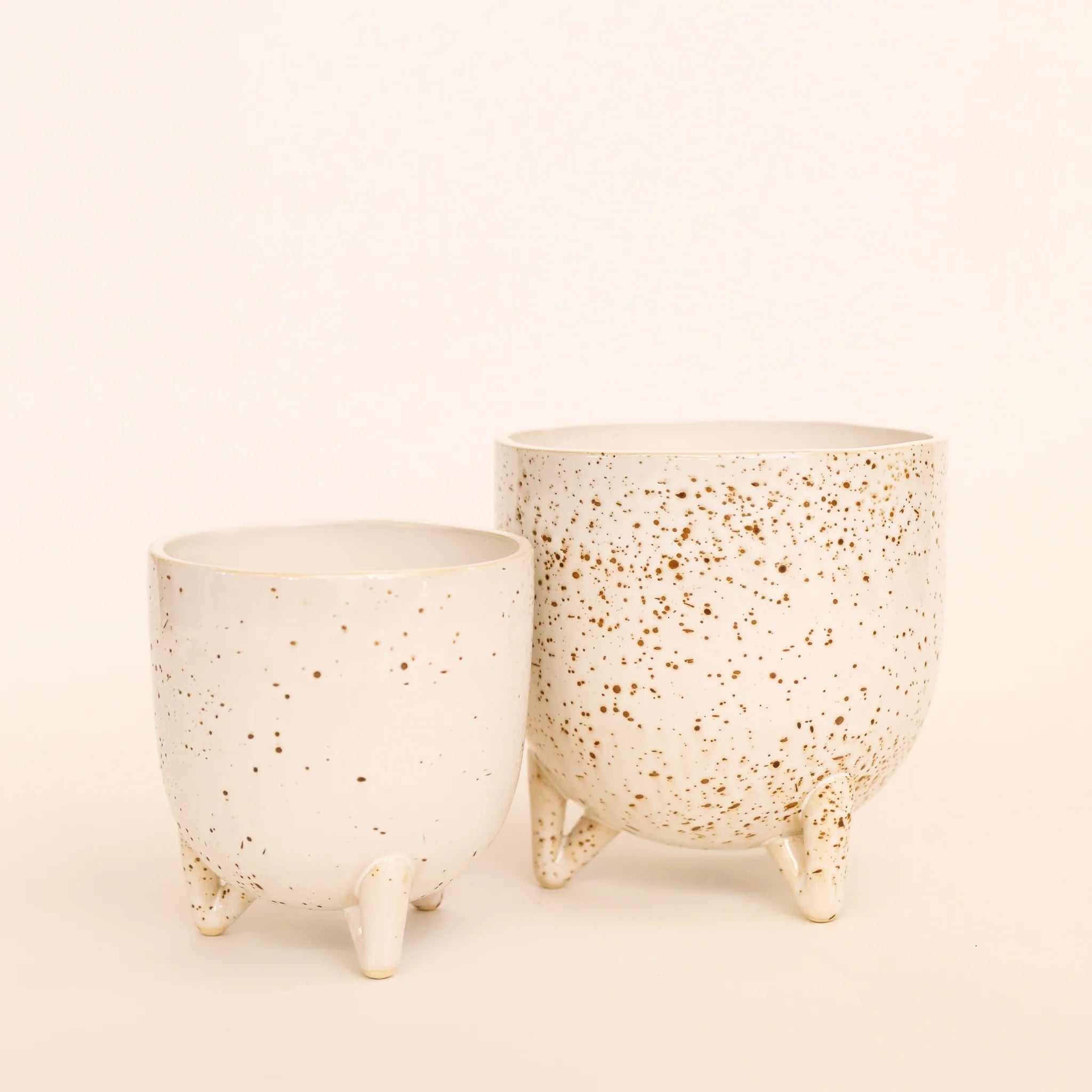 In front of a white background is a round footed pot. The pot is a cream color with dark brown specks. To the left is another pot just like the one on the right just a little smaller.