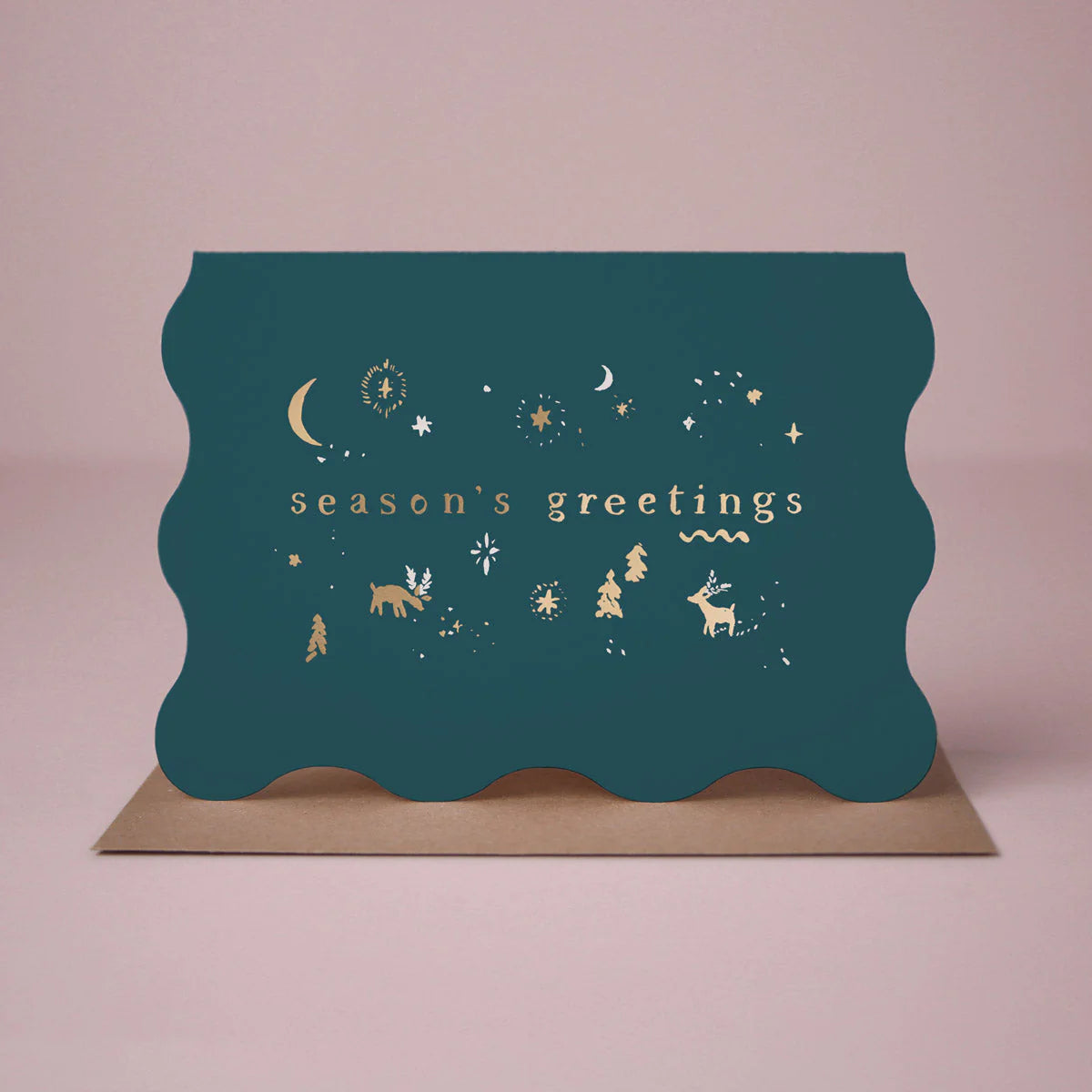 On a light pink background is a teal blue wavy edged greeting card with gold text and details that read, "season's greetings" along with a Kraft brown envelope.