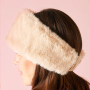 Side profile of a model wearing an oatmeal colored faux fur headband against a light pink background. 