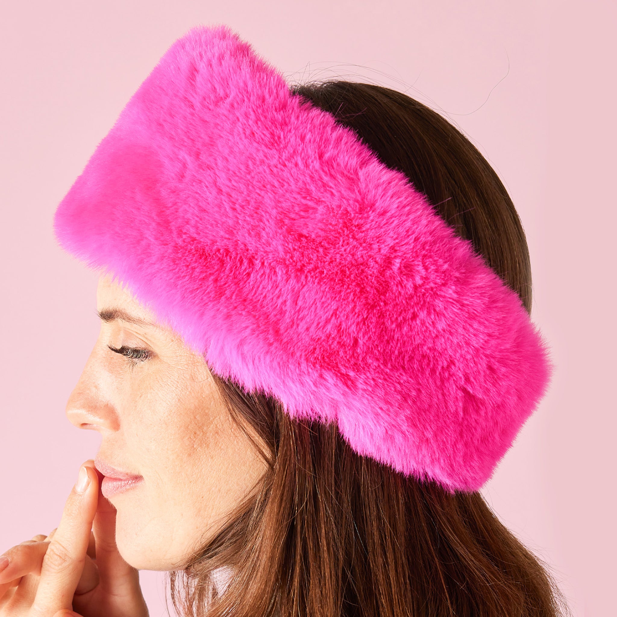 Side profile view of a model wearing a hot pink faux fur headband against a baby pink background. 