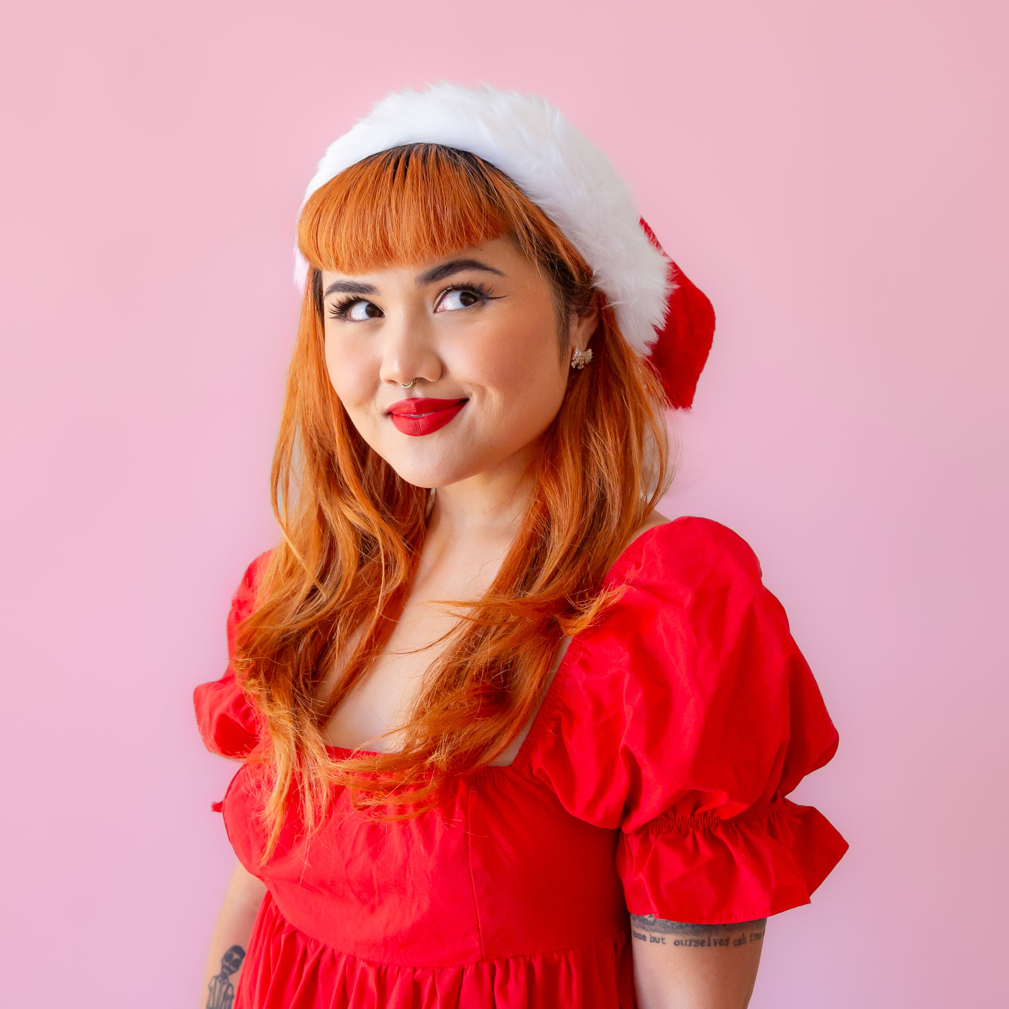 On a pink background is a model wearing a red Santa hat with white accents and faux fur.