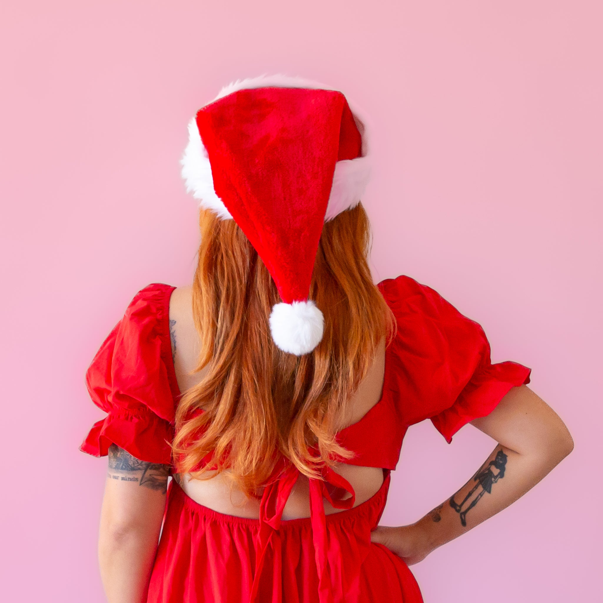 On a pink background is a model wearing a red Santa hat with white accents and faux fur.