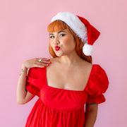 On a pink background is a model wearing a red Santa hat with white accents and faux fur.