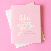 On a pink background is a light pink card with white text that reads, "We've Still Got It! Happy Anniversary".