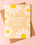 A tan card with white and yellow daisies all over the edges along with groovy text in the center that reads, "Welcome Little Babe" along with a Kraft brown envelope.