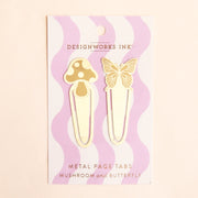 On a tan background is two gold book tabs one with a mushroom design and the other with a butterfly design and on a pink and white wavy cardboard packaging. 