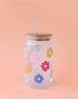A glass tumbler with multi colored daisies and a wooden lid and a glass straw.