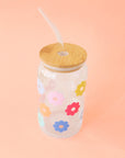 A glass tumbler with multi colored daisies and a wooden lid and a glass straw.