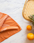 On a marble background is an orange kitchen towel with a white banana leaf design.