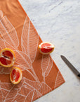 On a marble background is an orange kitchen towel with a white banana leaf design. 