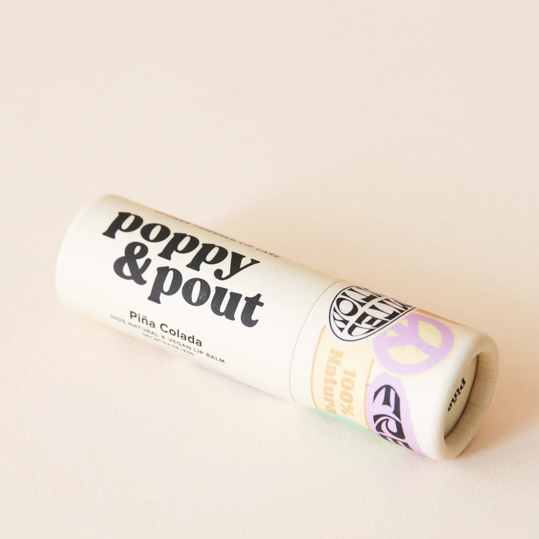 On a tan background is an ivory lip balm with black text that reads, &quot;poppy &amp; pout Pina colada&quot;. 