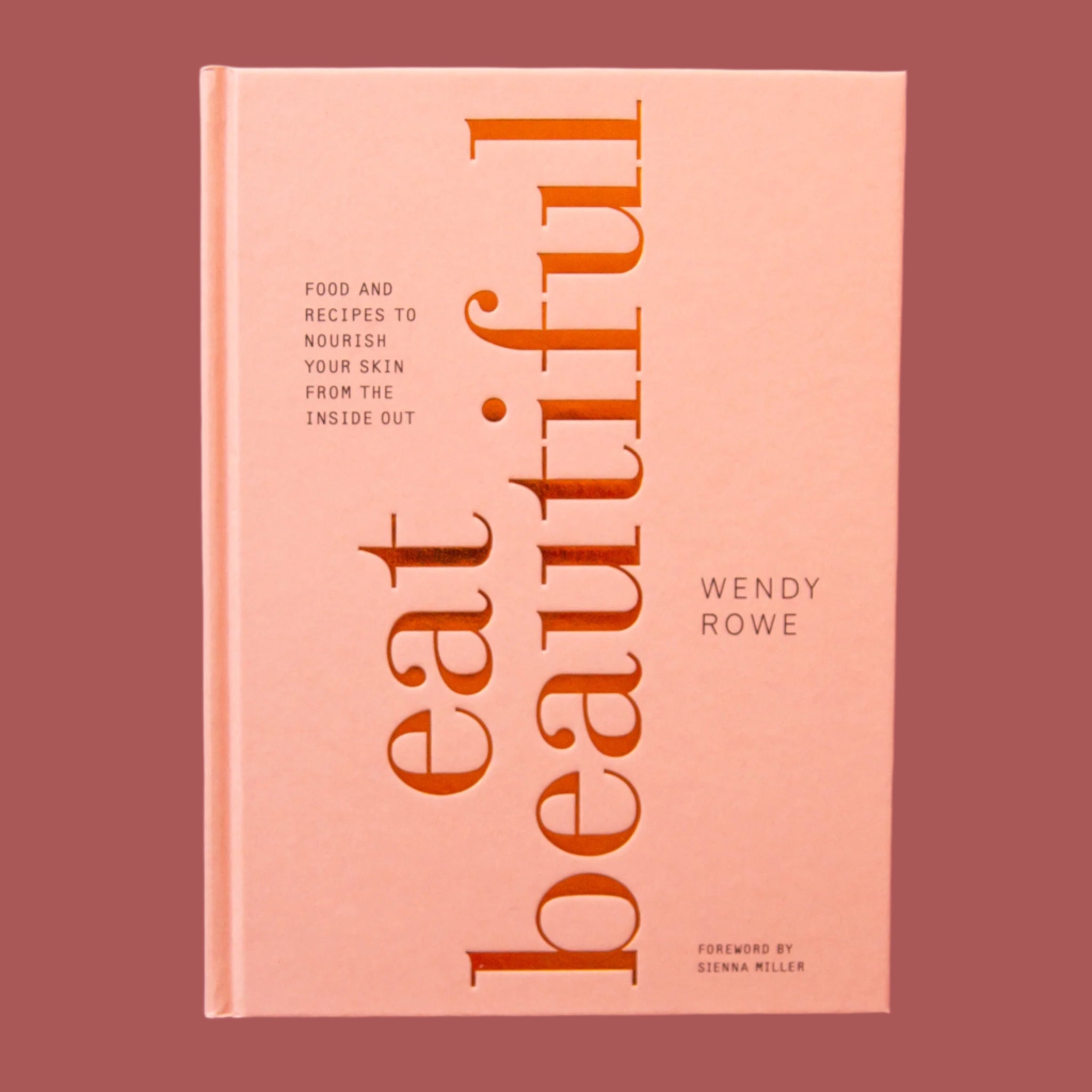 Cookbook with a soft pink hardcover that reads 'eat beautifully' in large gold foil lettering facing perpendicular to the cover. In smaller lettering reads 'food and recipes to nourish your skin from the inside out' in smaller lettering.