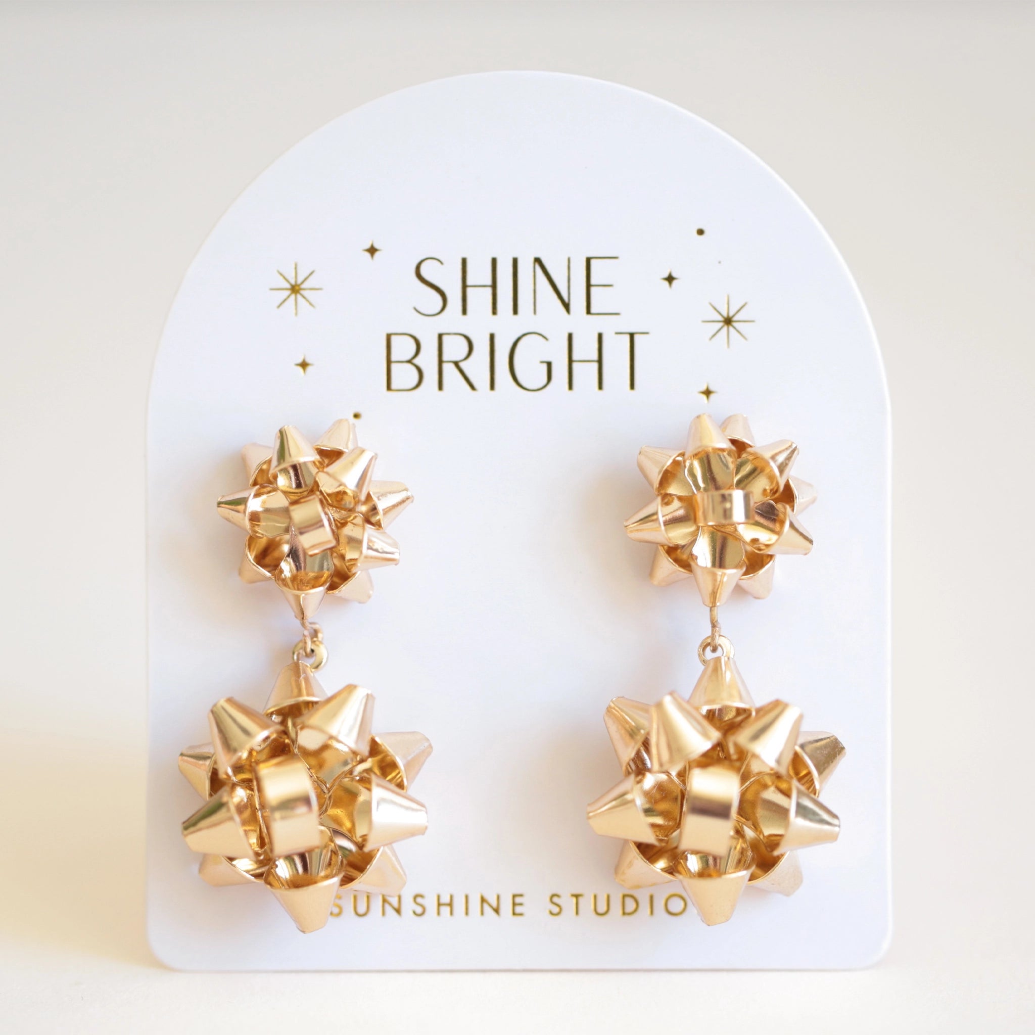 On an ivory background is a pair of gold holiday bow shaped earrings that have a smaller gold bow and a dangling larger bow attached to the bottom. The packaging reads, &quot;Shine Bright Sunshine Studios&quot;.