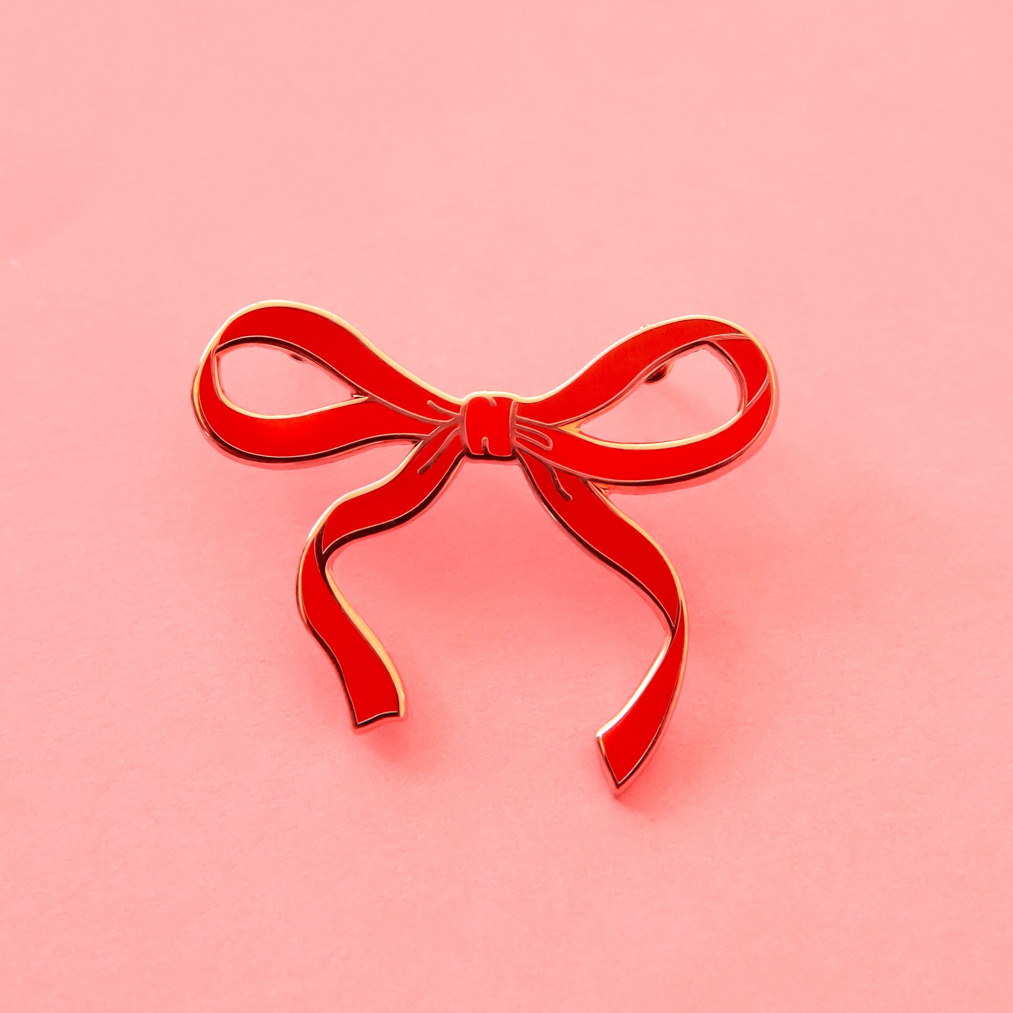 a beautiful red bow enamel pin lies on a pink background.  the pin has a gold outline casing.