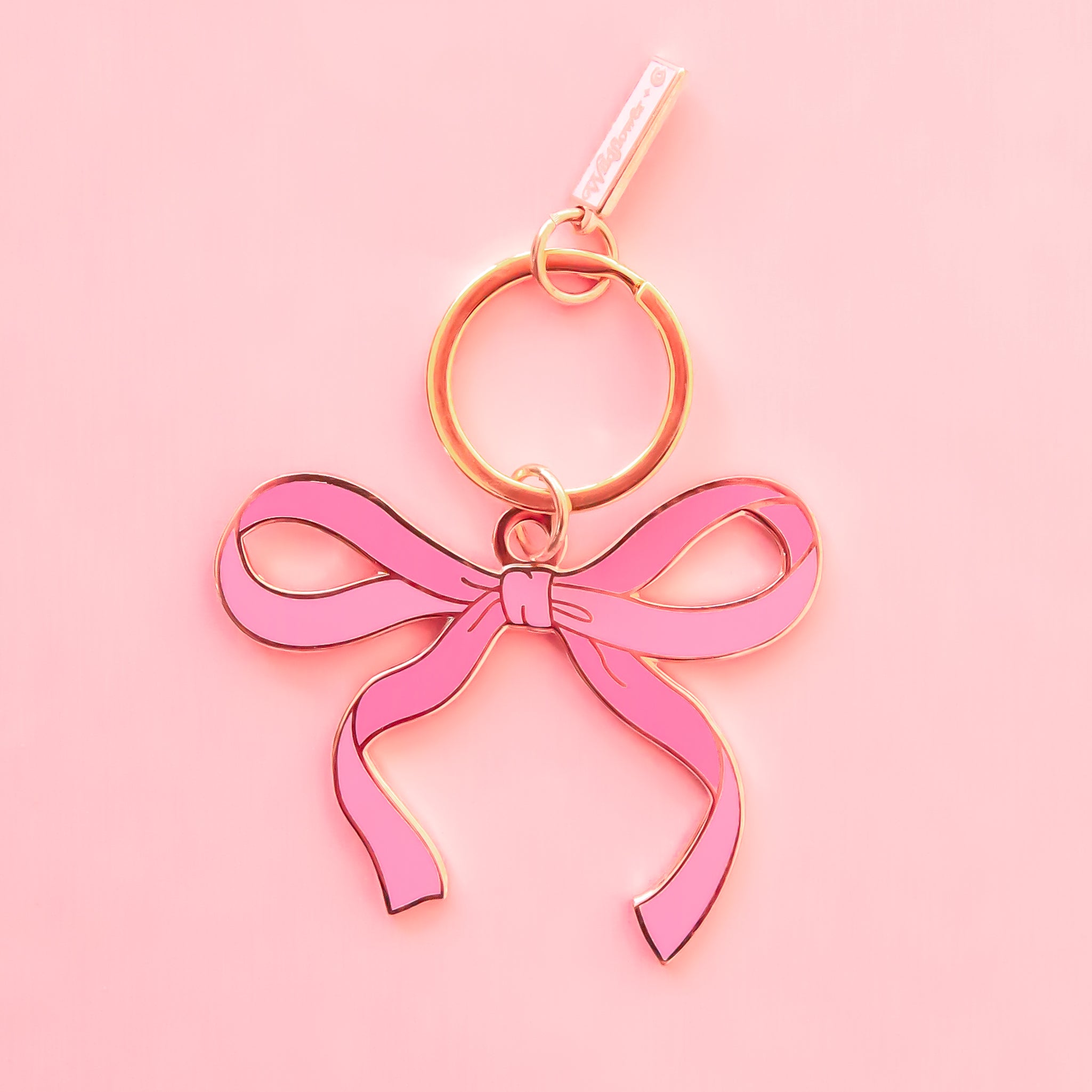 a pink enamel bow shaped keychain sits on top of a pink background. 