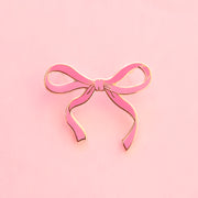 A pink enamel bow pin sits on top of a pink background.  There is a gold brass border around the enamel lapel pin.