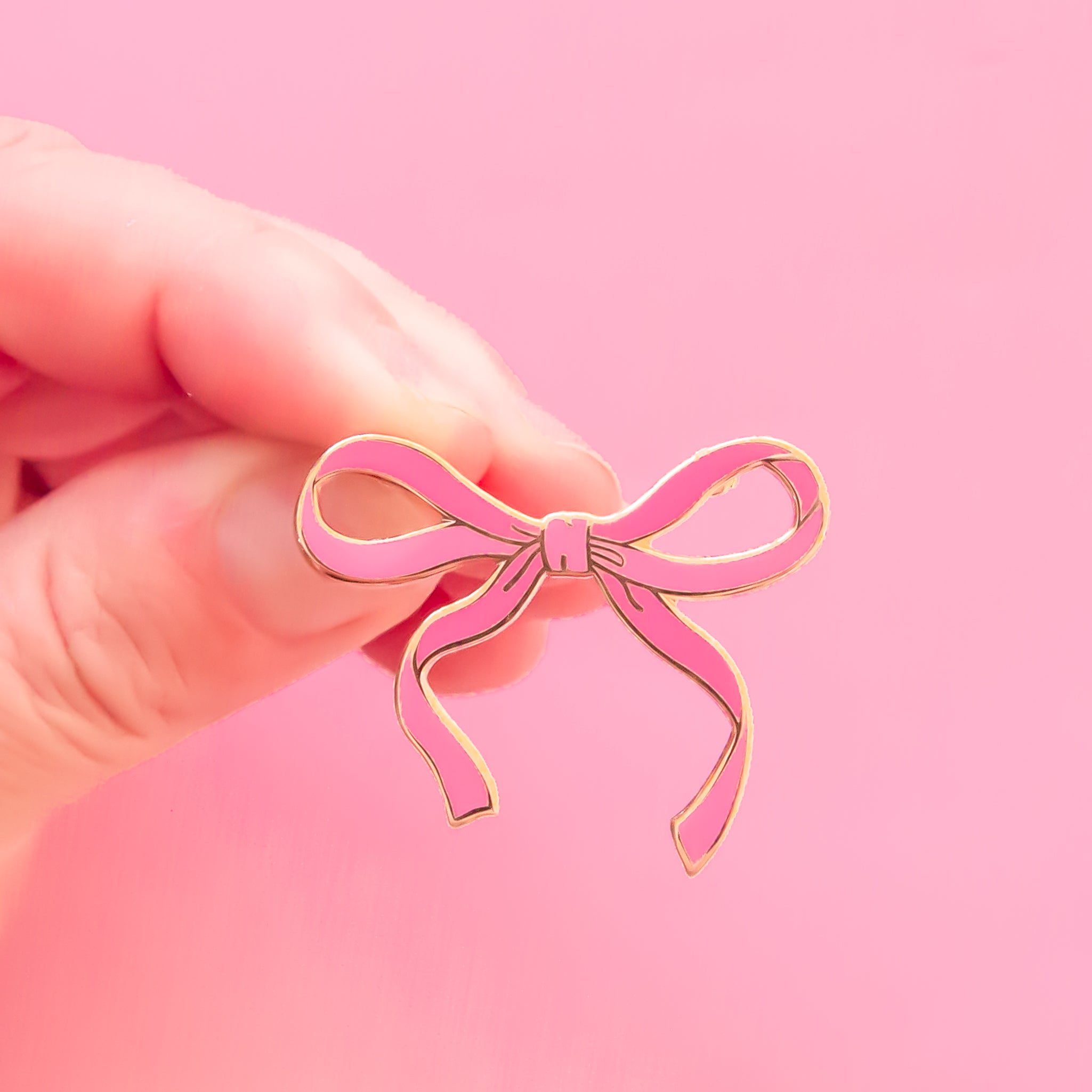 Fingers coming into frame holding a pink bow lapel pin with gold border. 