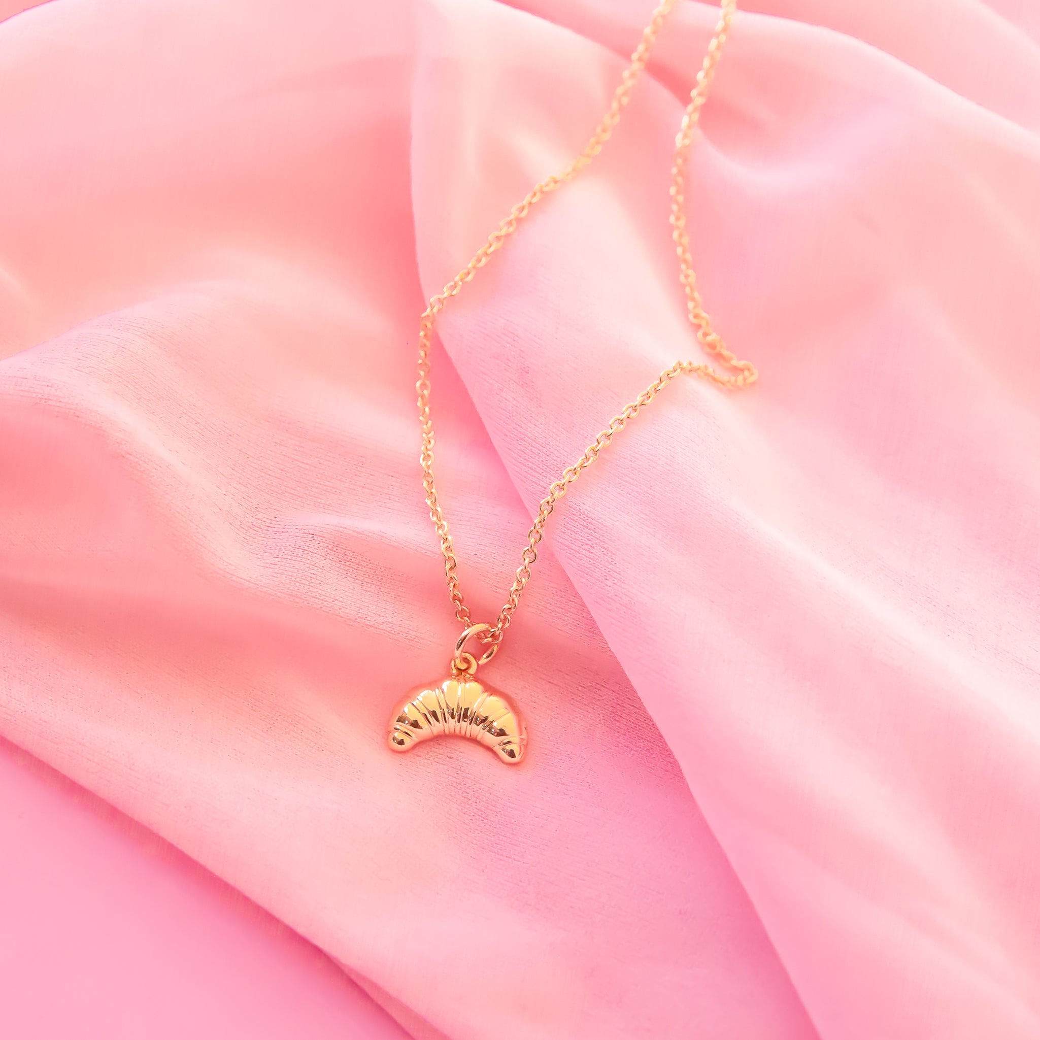 gold croissant shaped charm hangs on a gold necklace chain.  the backrgound of the image is a soft pink chiffon fabric.