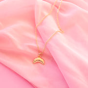 gold croissant shaped charm hangs on a gold necklace chain.  the backrgound of the image is a soft pink chiffon fabric.