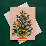 A cream colored card sits against a dark green background. On the front of the card there is a drawing of a dark green Christmas tree. There are yellow and red Christmas lights up and down the tree. There are green, red and pink ornaments spread out all over the tree. Silver tinsel is hanging on the end of each tree branch. At the bottom is a red tree skirt. At the top is a sliver star. Above the tree in silver script it reads ‘season's greetings.’ The card is accompanied by a white envelope.