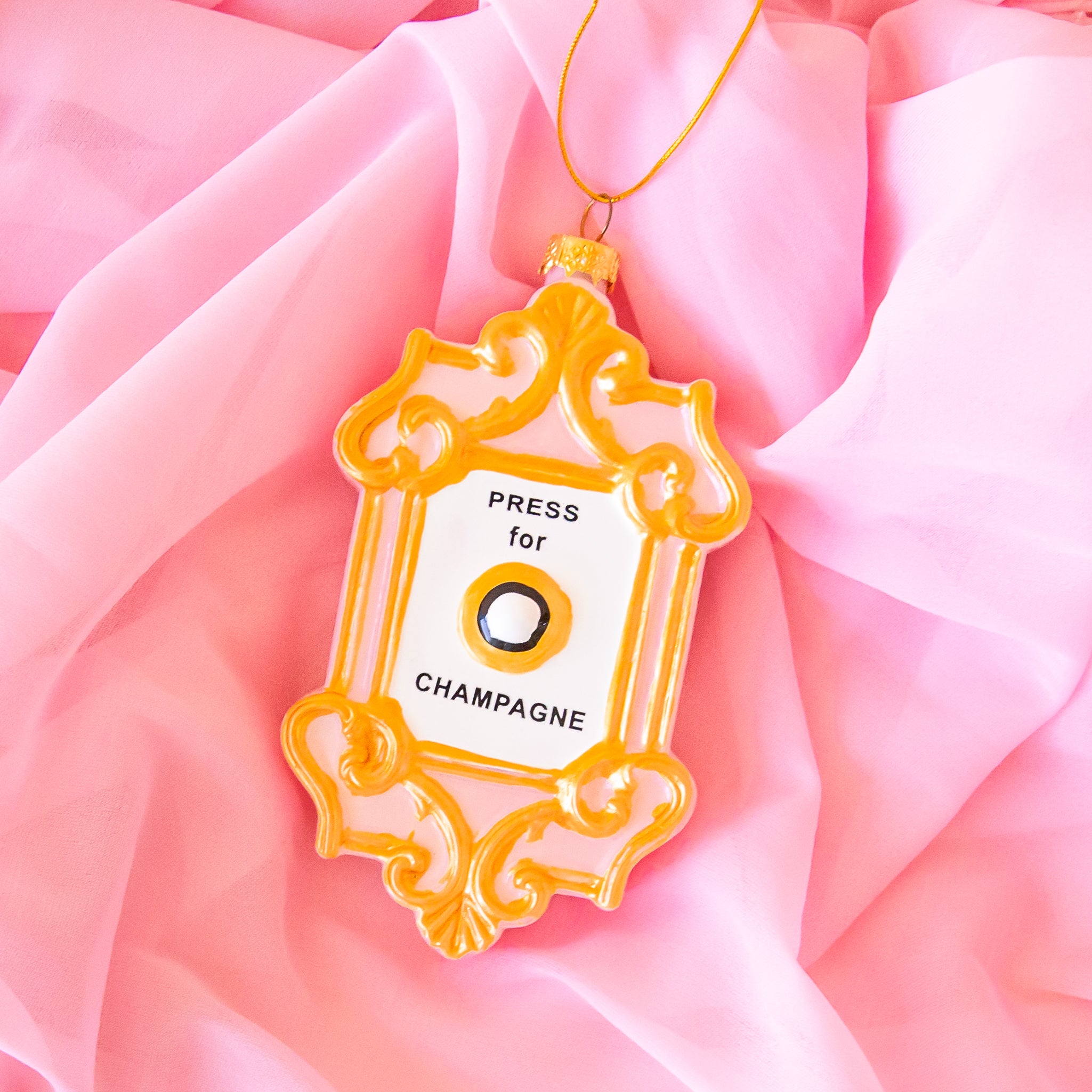 On a pink chiffon fabric background is an ornament in the shape of a champagne button with gold details.  It reads, &quot;press for champagne&quot;.