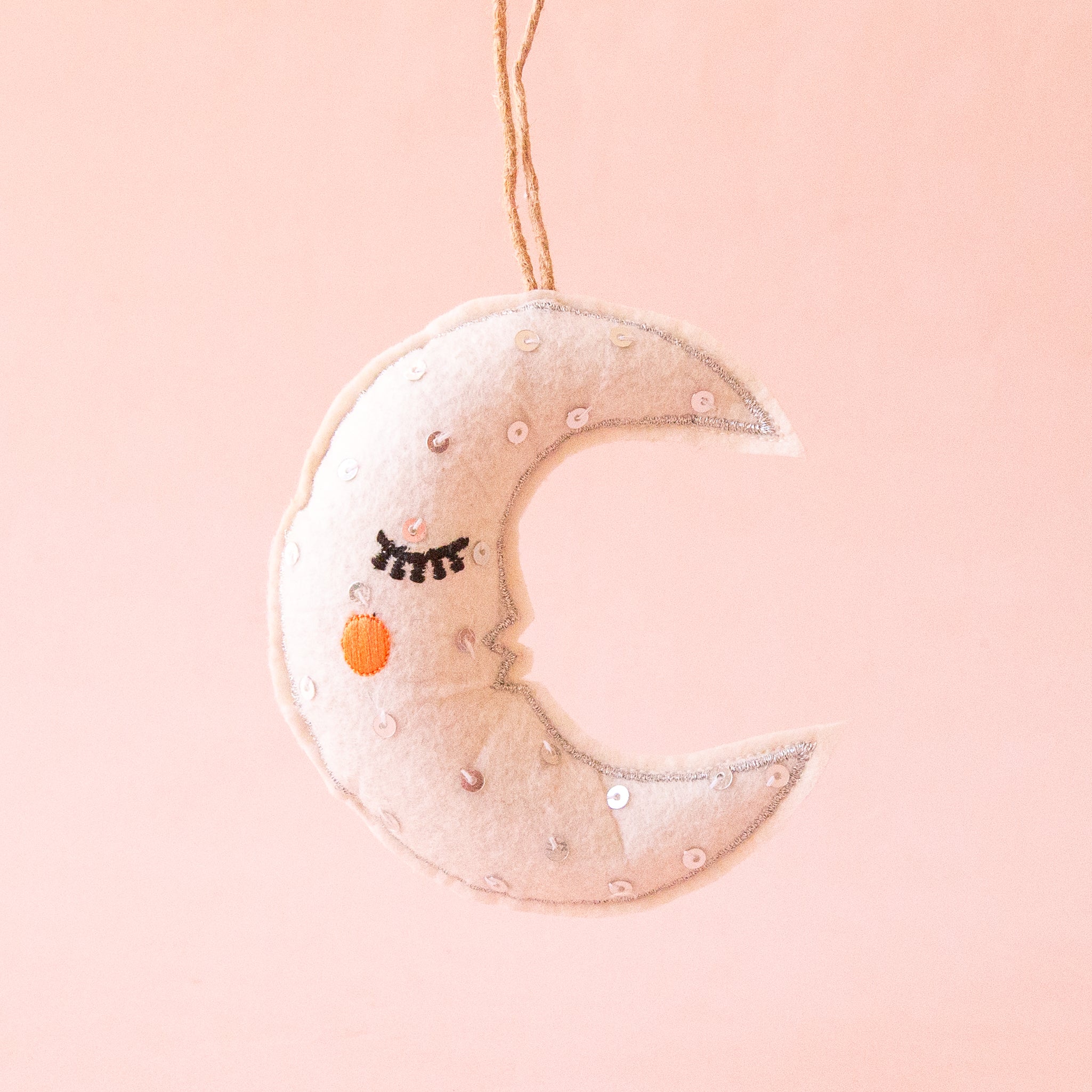 Photo is a felted moon ornament hanging on a jute piece of string.  The moon is white with black eyelashes and an orange circle cheek.  The moon also has silver sequins sewn onto it.