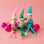 Three holiday elves sit on a pink background on top of hot pink glass ball ornaments.  The elves are in a pink, green and turquoise elf outfit with tall hats and tinsel neck details.