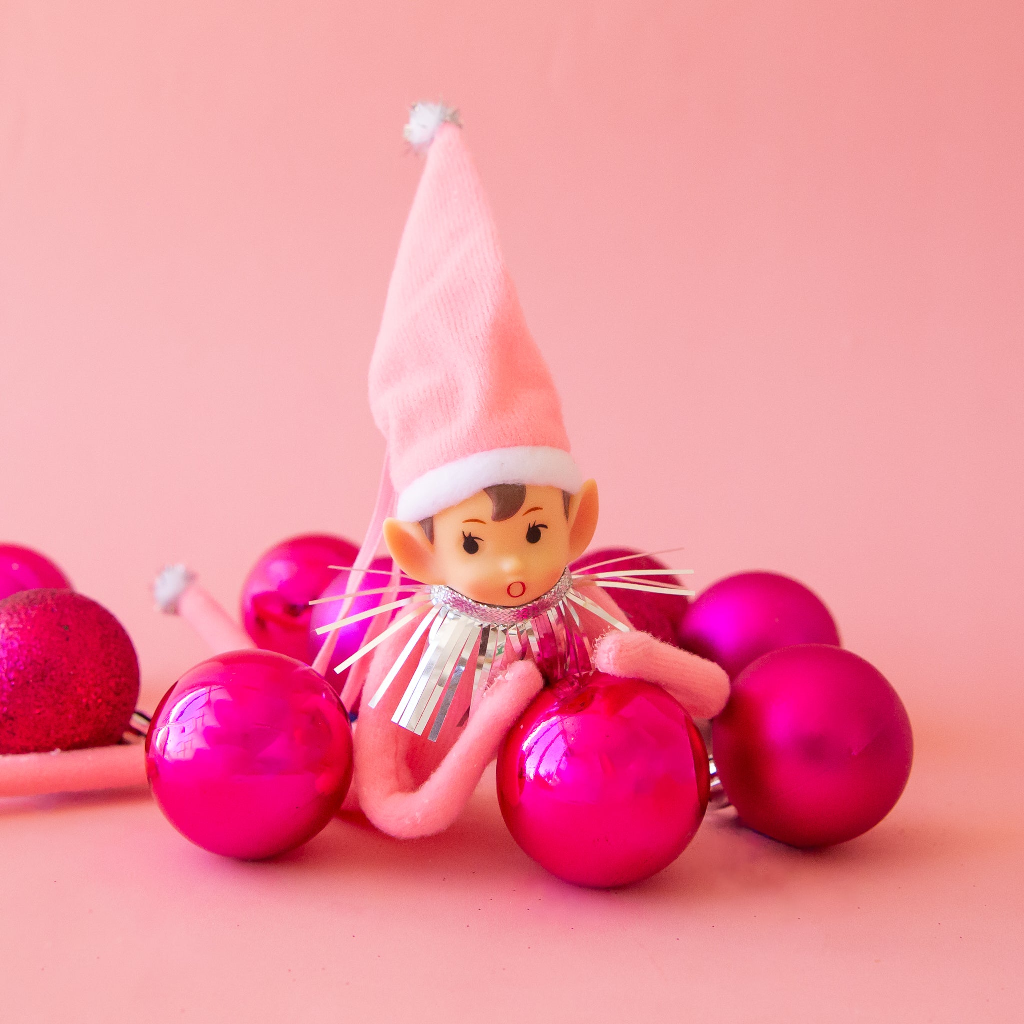 An adorable pink elf wearing a pink tall hat with silver tinsel around it's neck rests on hot pink christmas round ball ornaments. 