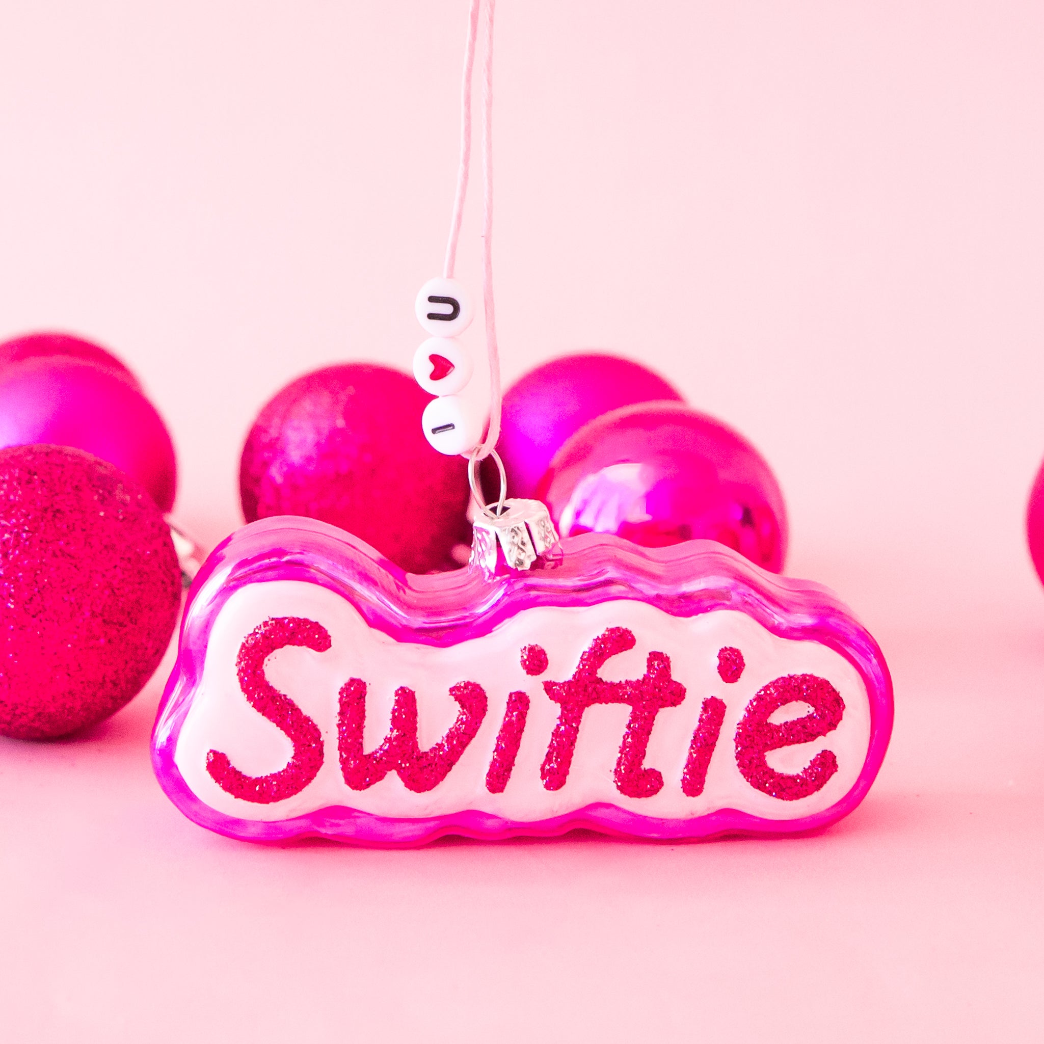 pink ornament that reads "swiftie" in red glitter writing. background is pink with silver tinsel and confetti.