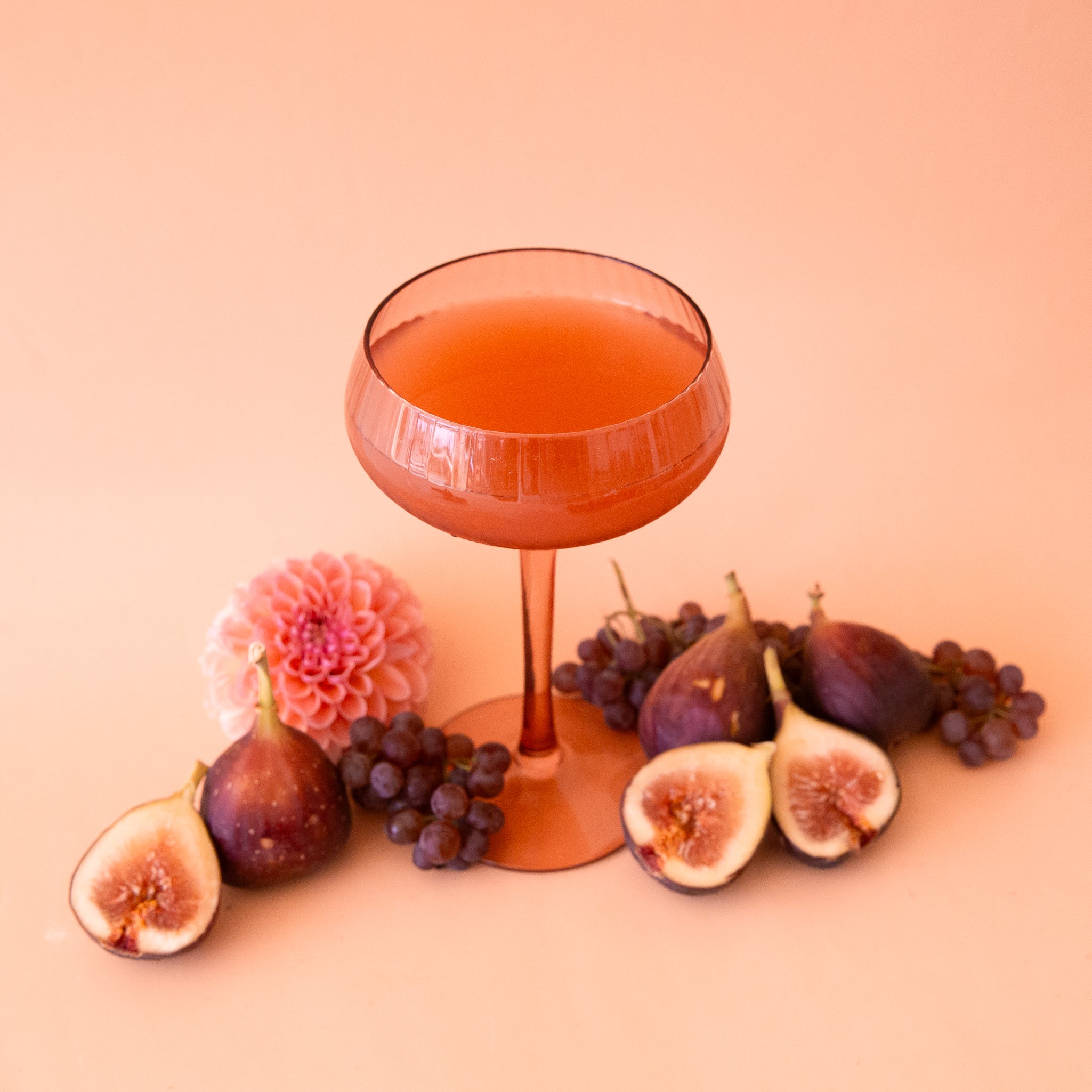 A light pink ribbed coupe cocktail glass surrounded by figs and grapes not included with purchase. 