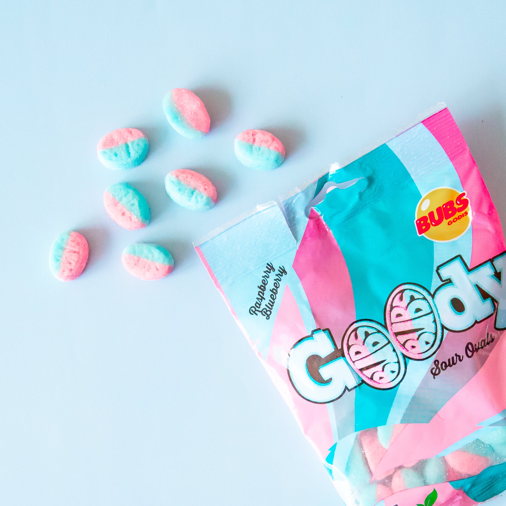 A bag of blue and pink candy spills onto a blue background.  The plastic candy bag reads &quot;goody sour ovals&quot;.  The flavor listed on the bag is Rasberry Blueberry. The candies spilling out are pink and blue striped.