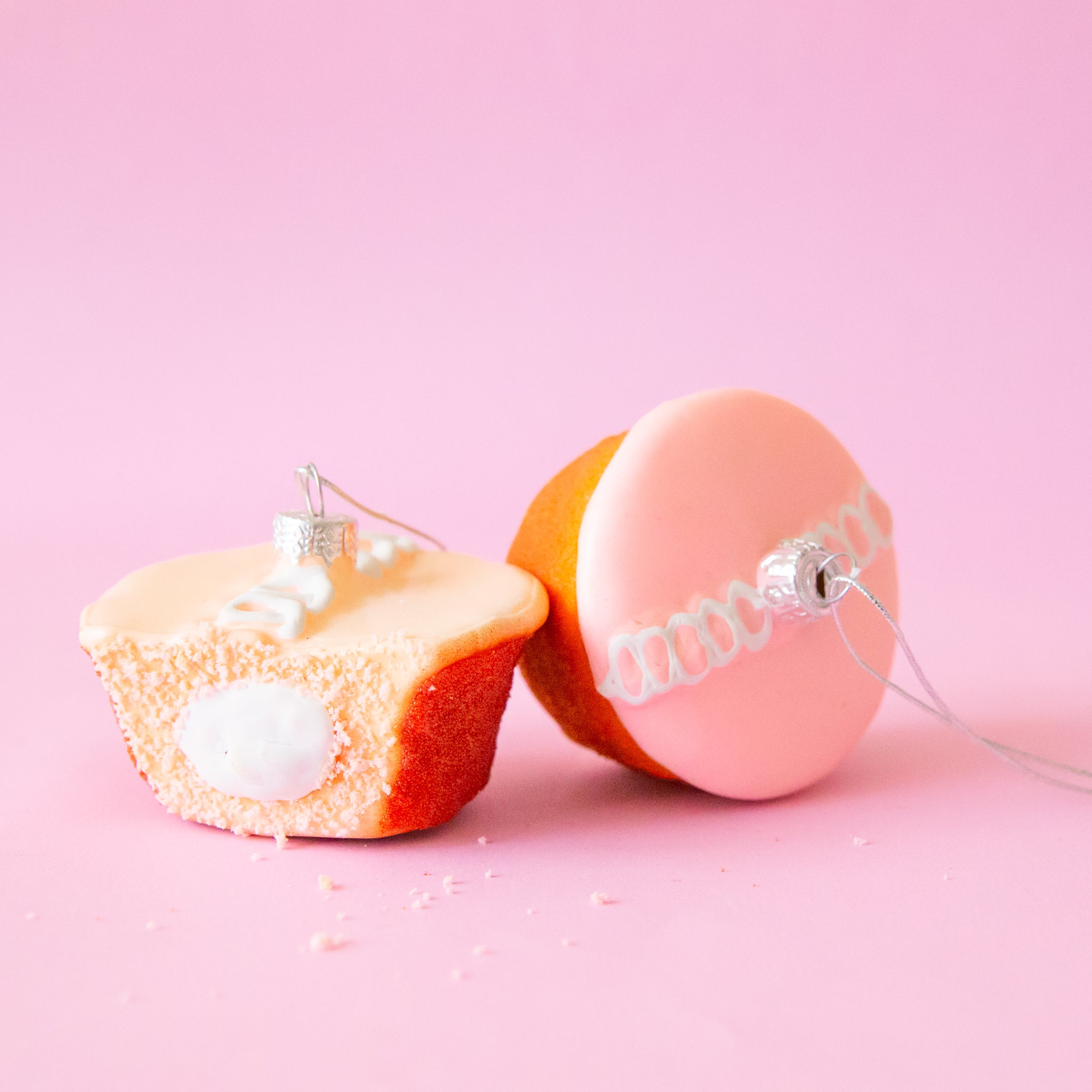 Two pink cupcake ornaments sit side by side.  The one on the left has a bite taken out of it exposing white creme filling.  The second cupcake is on it's side and top has white circle detail icing.