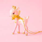 A small pink deer with gold glitter spots and feet stands on a pink ground.  The deer has a big gold bow around it's neck and a gold jingle bell.