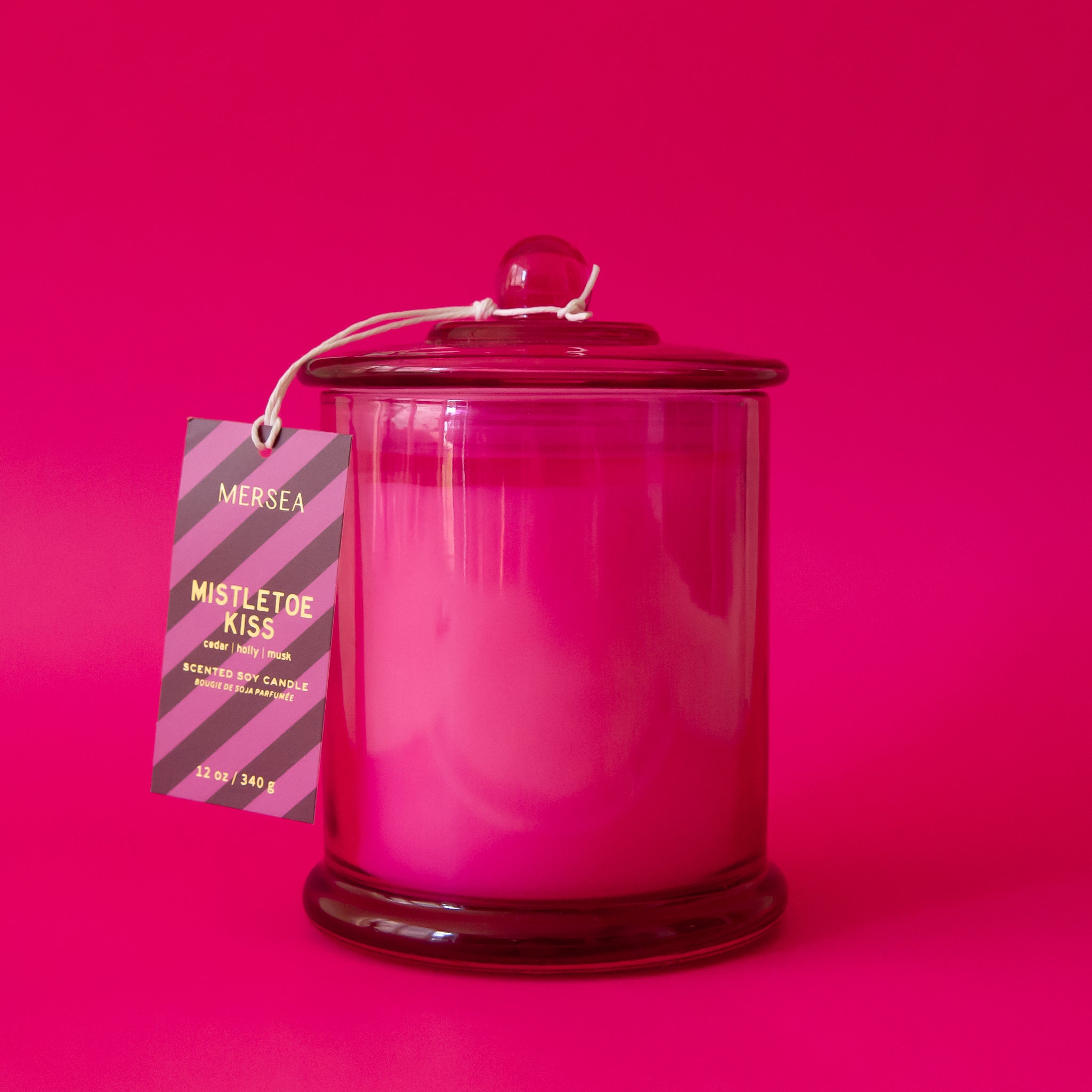 A beautiful hot pink glass cylinder shaped candle sits in the middle of image.  The lid has a little round knob with a paper tag hanging off that reads &quot;Mistletoe Kiss&quot;.  The label has purple and pink stripes and hangs from a white string tied around the round knob.  The background of the image is also hot pink.