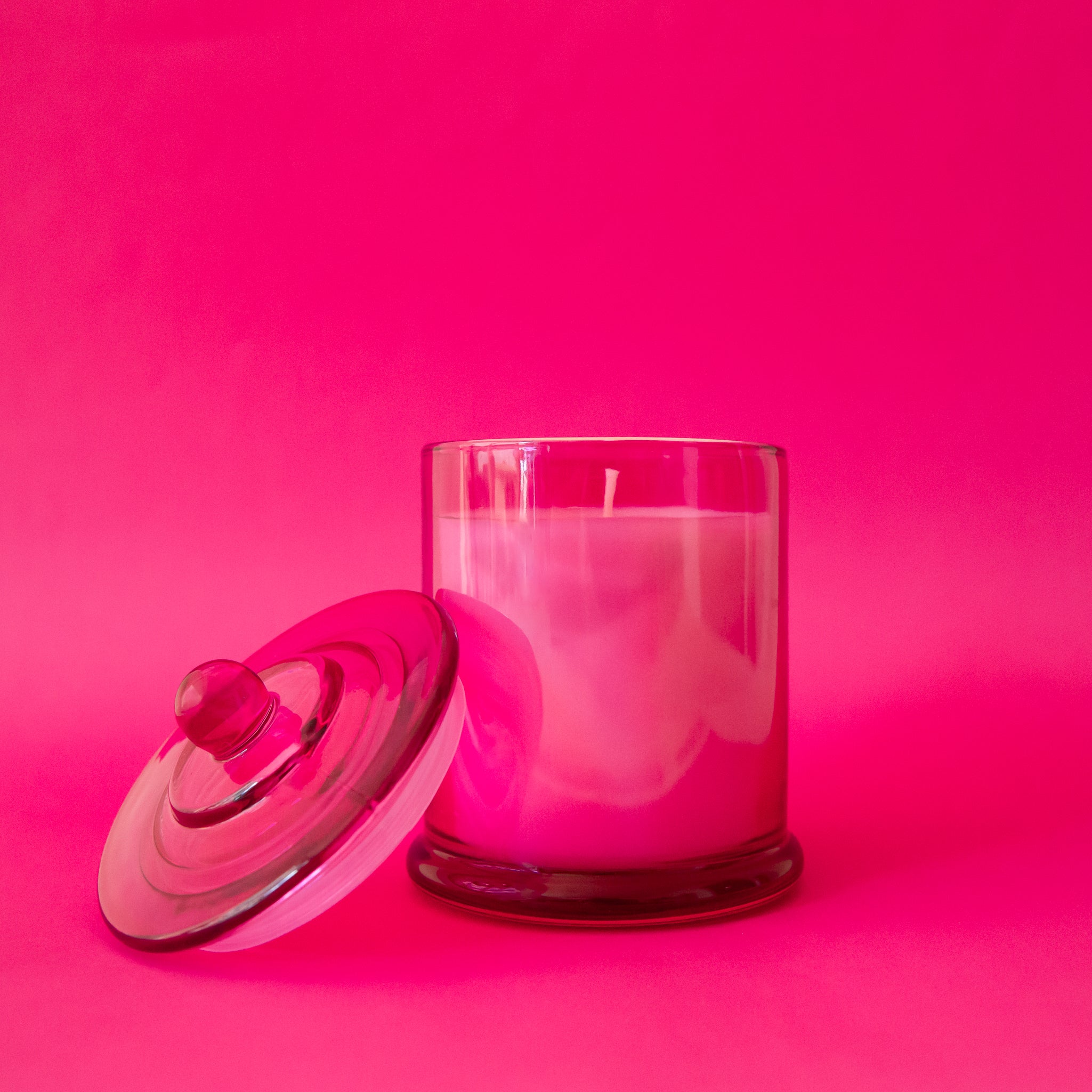 A beautiful hot pink glass cylinder shaped candle sits in the middle of the page.  The glass lid with a round knob sits on the left hand side at an angle resting on the candle.  You can see the white wick through the glass.  The background of the image is also a beautiful hot pink color.