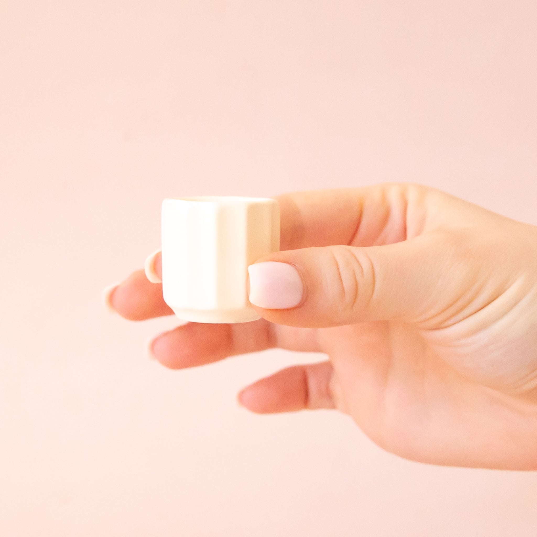 An itty bitty pot about the size of thimble that is a white color sits in the middle of a page.  The background is a soft pink color.