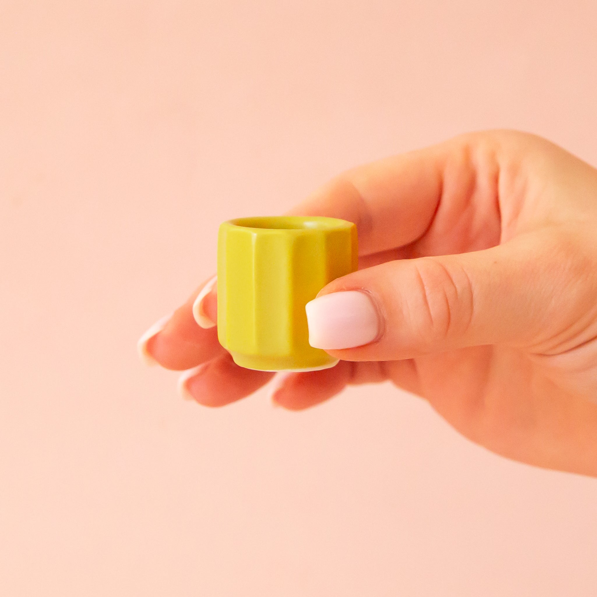 A hand comes into frame on the right hand side of image with soft pink nails.  The hand is holding an itty bitty pot about the size of thimble that is a chartreuse color.  The background is all  a soft white color.