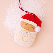 On a light pink background is a light brown Santa face ornament made of felt with a red Santa cap on. 