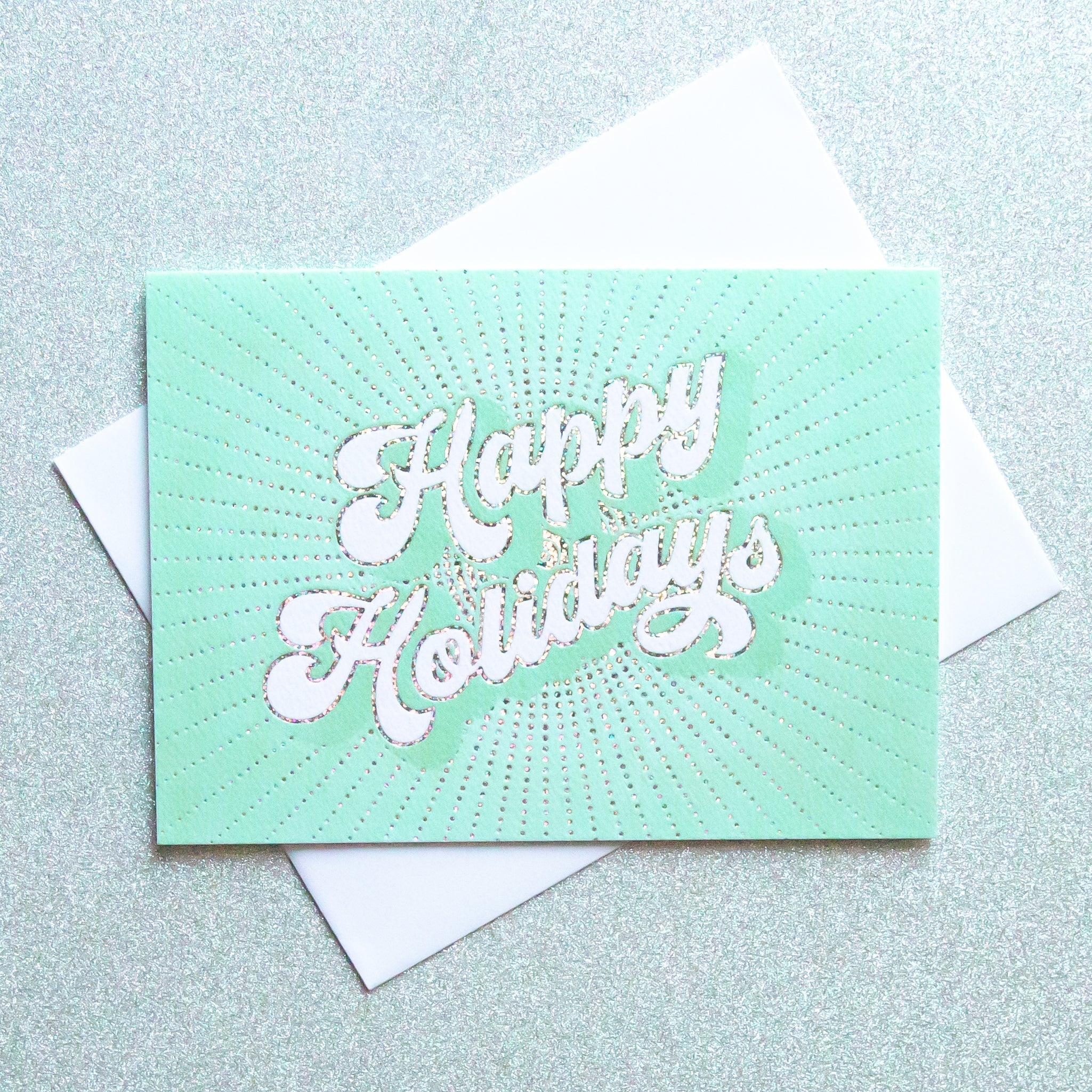 On an ivory background is a teal holiday greeting card with white text that reads, "Happy Holidays" along with a coordinating white envelope.