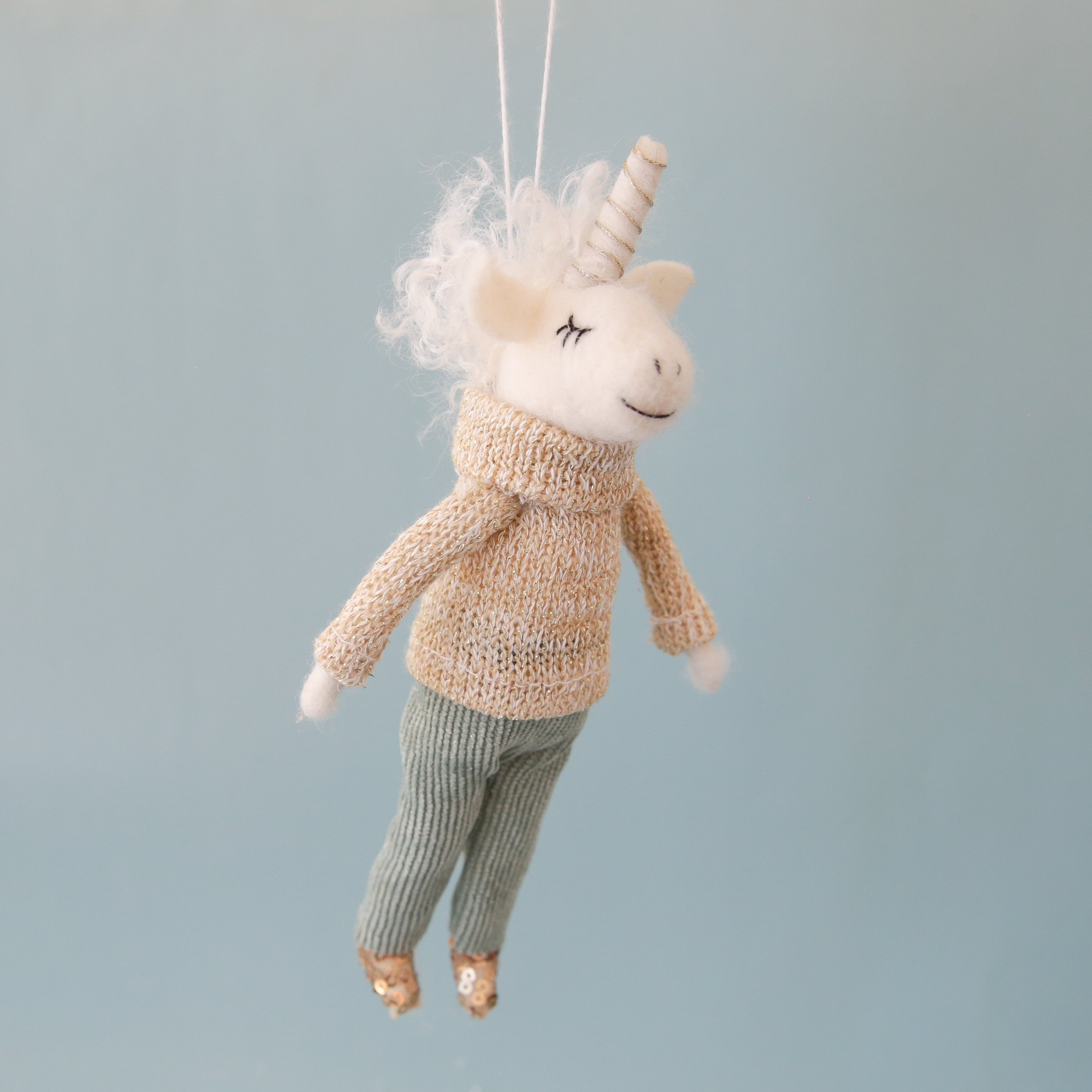 On a aqua blue background is a white felt unicorn ornament with a knit sweater on and corduroy pants and sequin shoes