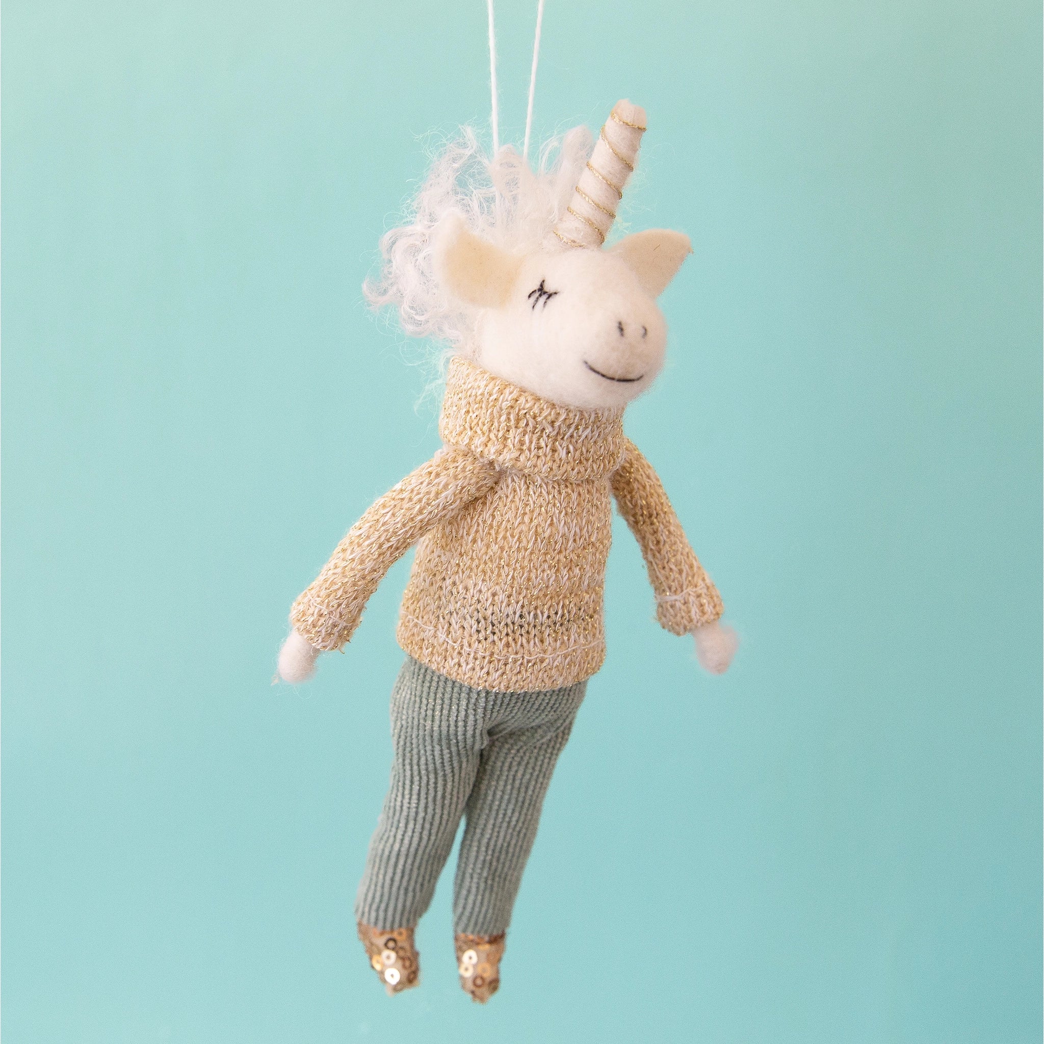 On a aqua blue background is a white felt unicorn ornament with a knit sweater on and corduroy pants and sequin shoes. 