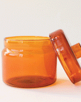 A round glass amber jar with a coordinating glass lid that has a round, flat knob on top.