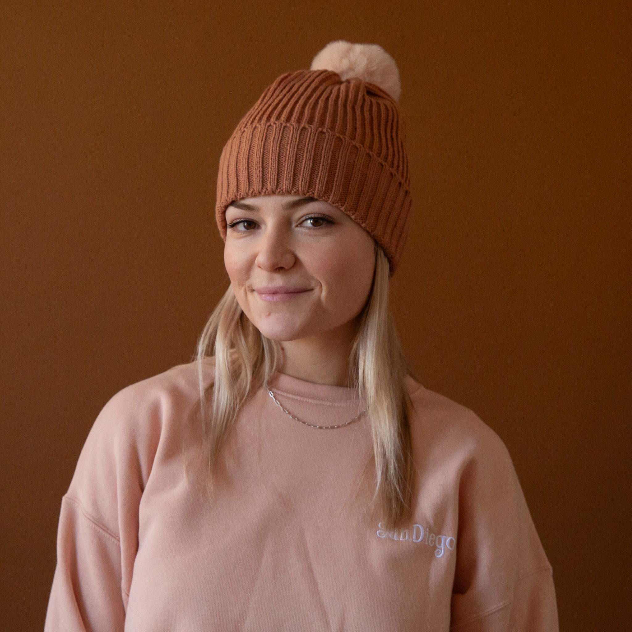 A model wearing a light brown beanie with a neutral pom at the top. 