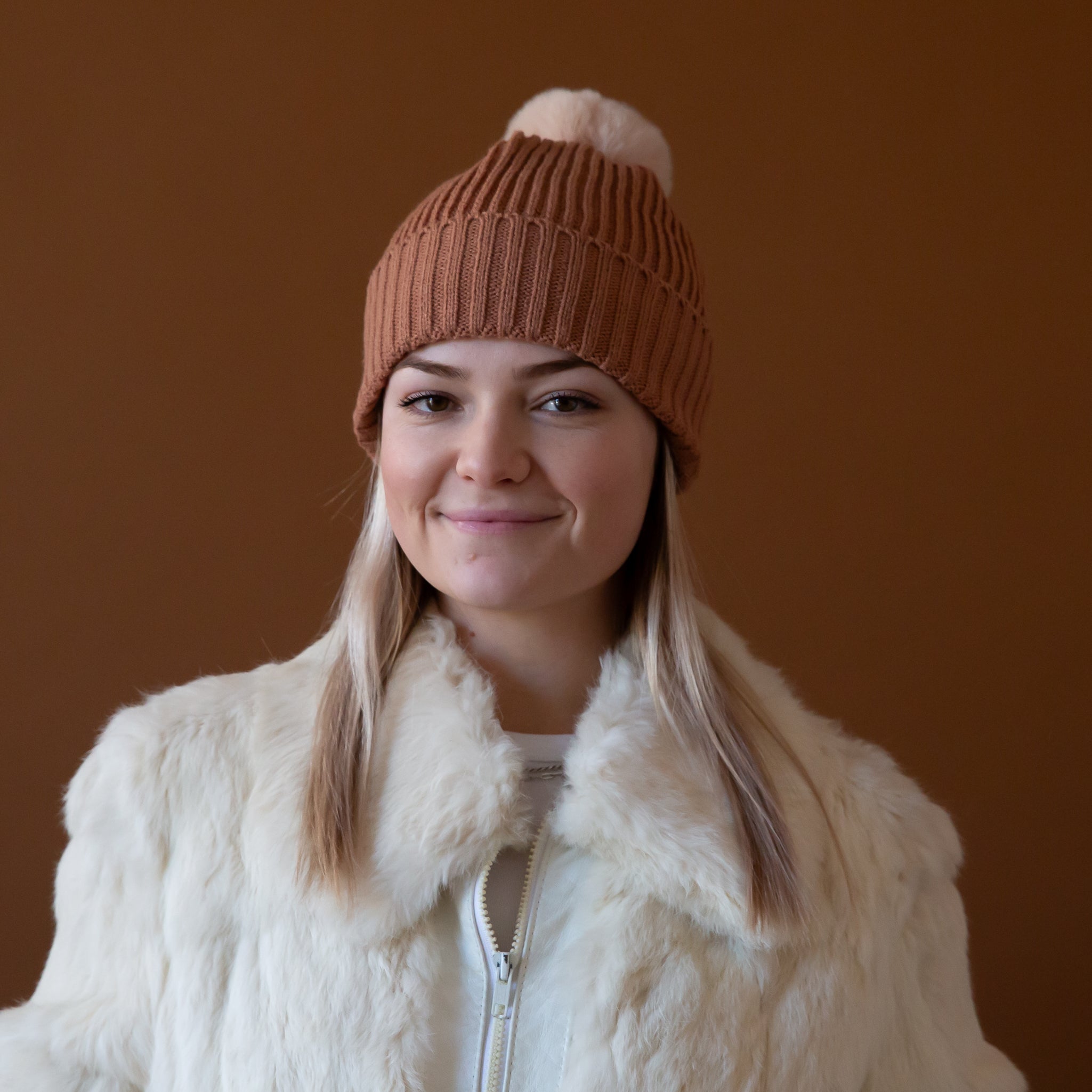 A model wearing a light brown beanie with a neutral pom at the top. 