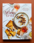 Hard covered cookbook titled 'Wine Style: Discover the wines you will love through 50 simple recipes' in red lettering. Book cover features brittle salted crackers and dip along side wine-filled cocktail glasses against white modern tiled.