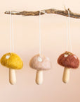 Three felt mushroom and wood ornaments hung on a stick. From left to right there is a yellow, neutral tan and then cocoa brown shade. 