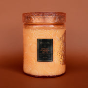 On a burnt orange background is a large decorative jarred candle in an orange shade with a matching lid and black label in the center that reads, "Voluspa Spiced Pumpkin Latte".